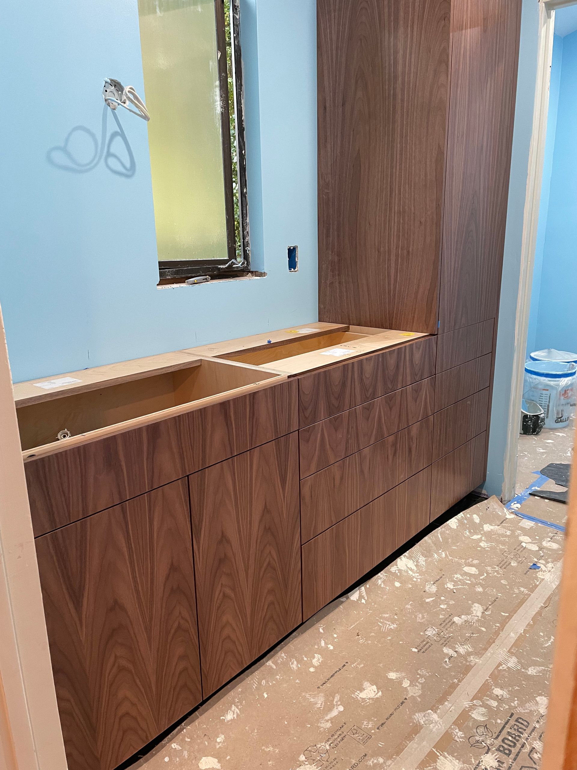 Wood vanity cabinets under construction beside a mirror in a light blue bathroom, with tools and debris on the floor