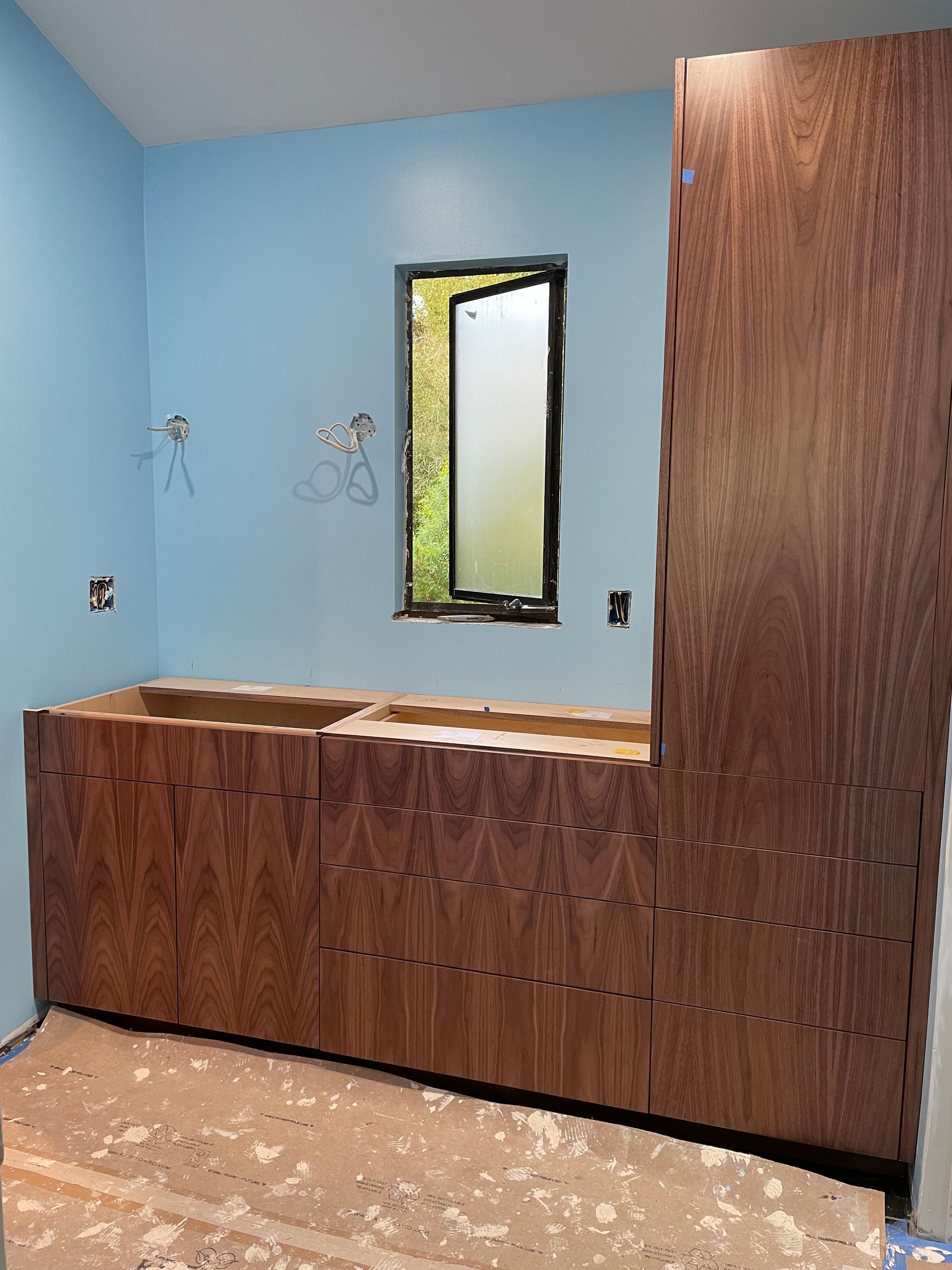 Bathroom vanity under installation with blue walls, wood cabinets, and a narrow vertical window.