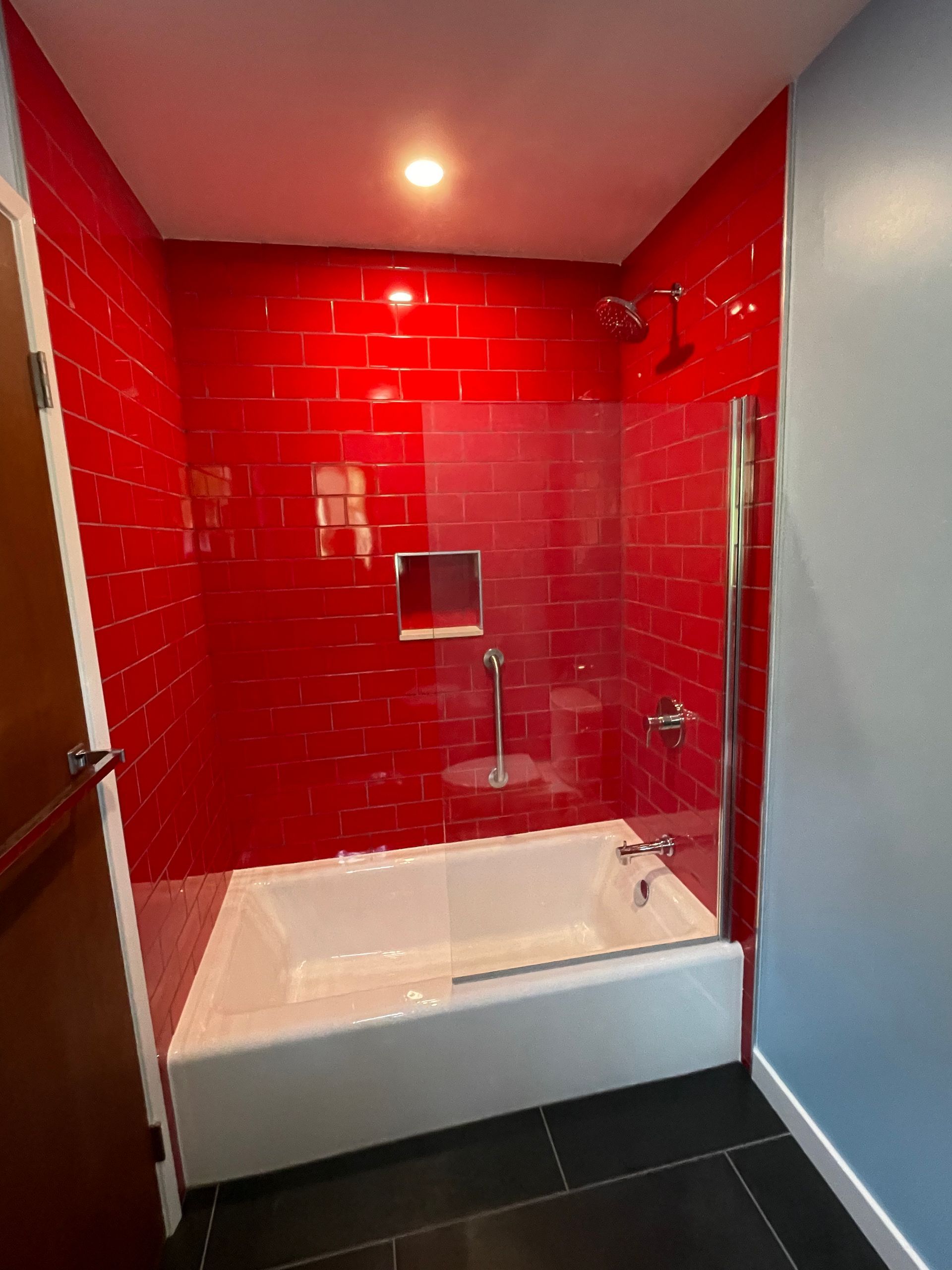 Red-tiled shower alcove with white bathtub, glass panel, and dark floor tiles