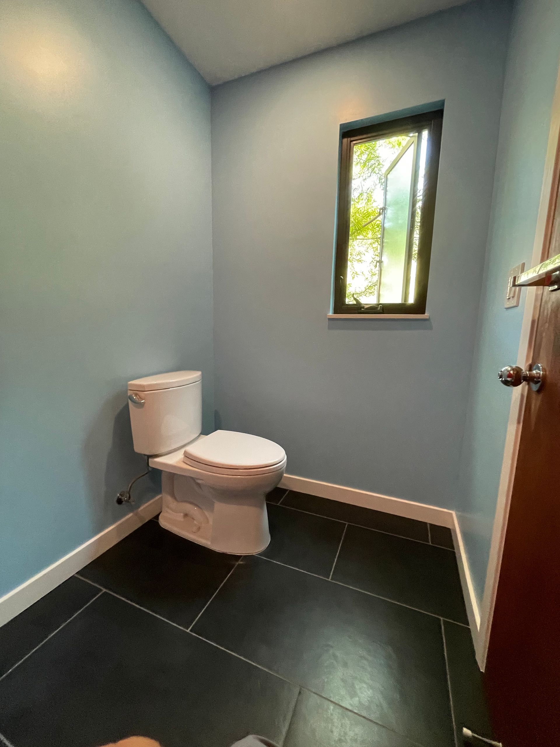 Small bathroom with light blue walls, a white toilet, black tile floor, and a narrow window.
