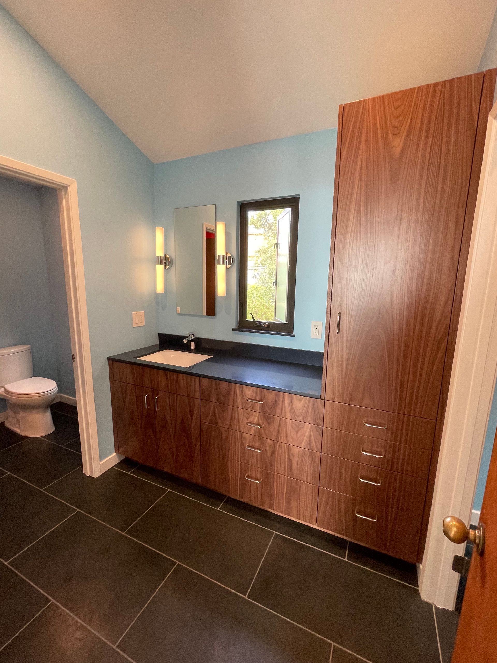 Modern bathroom vanity with wood cabinets, black countertop, mirror, and toilet in a blue-walled room.