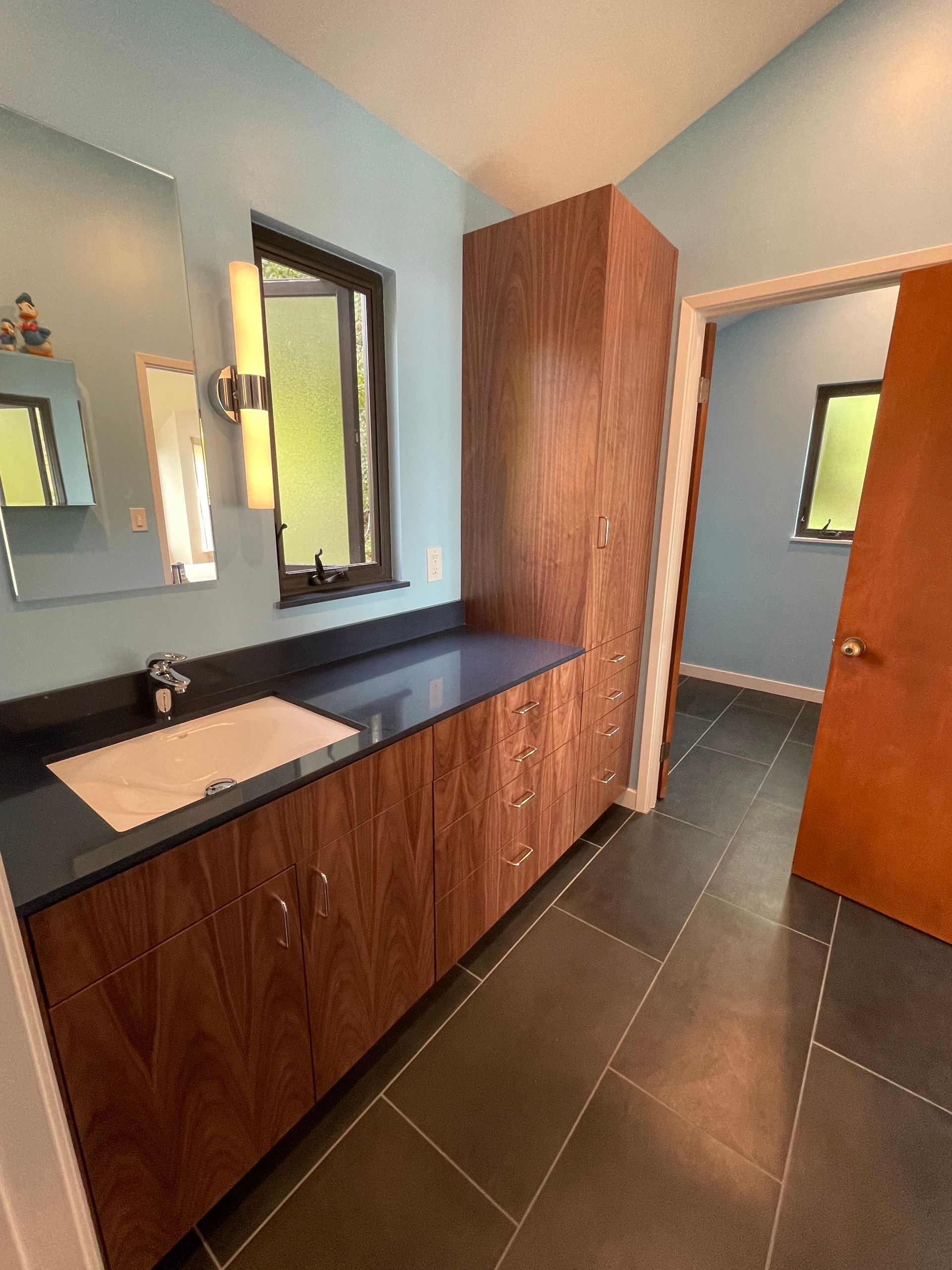 Modern bathroom with wood vanity, black countertop, mirror, and open door to a tiled room.