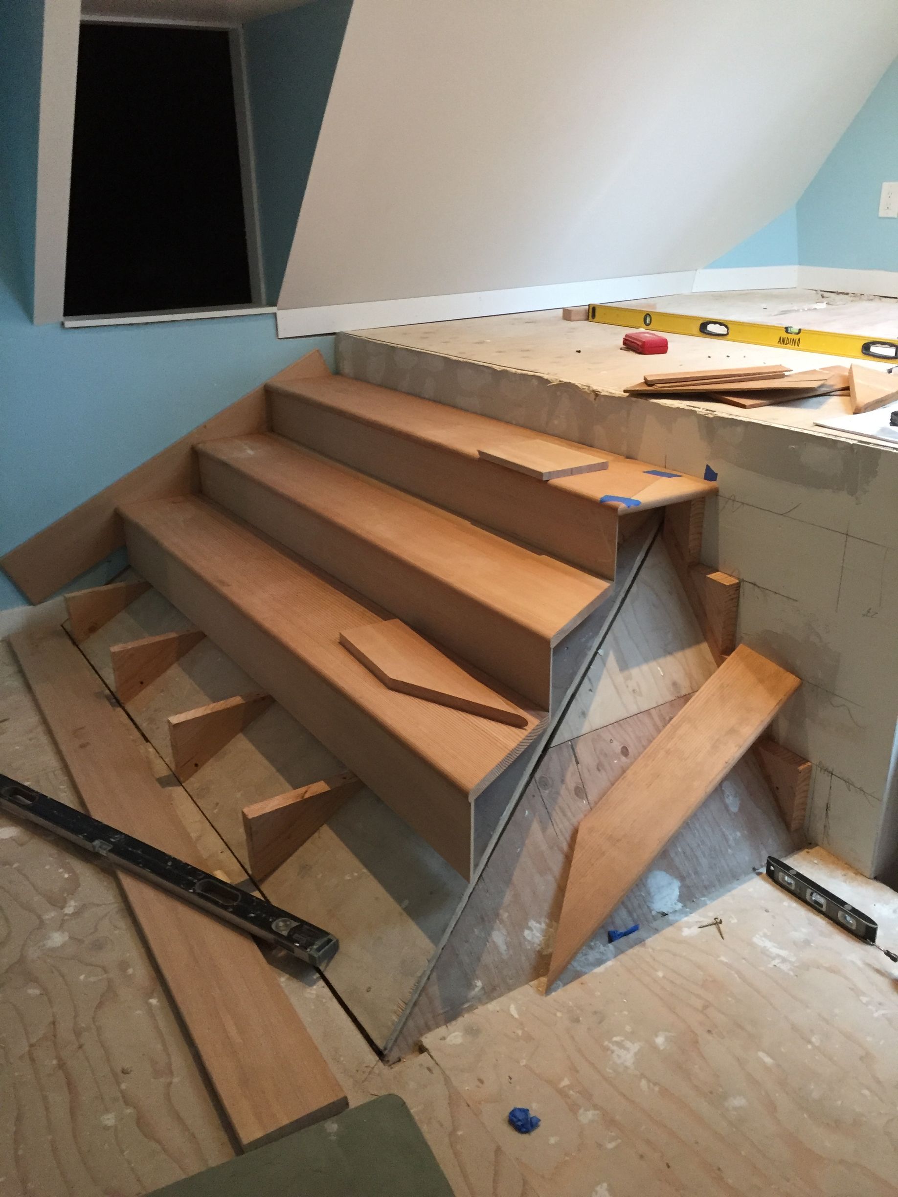 Wooden staircase under construction in a small attic room with unfinished walls and tools nearby