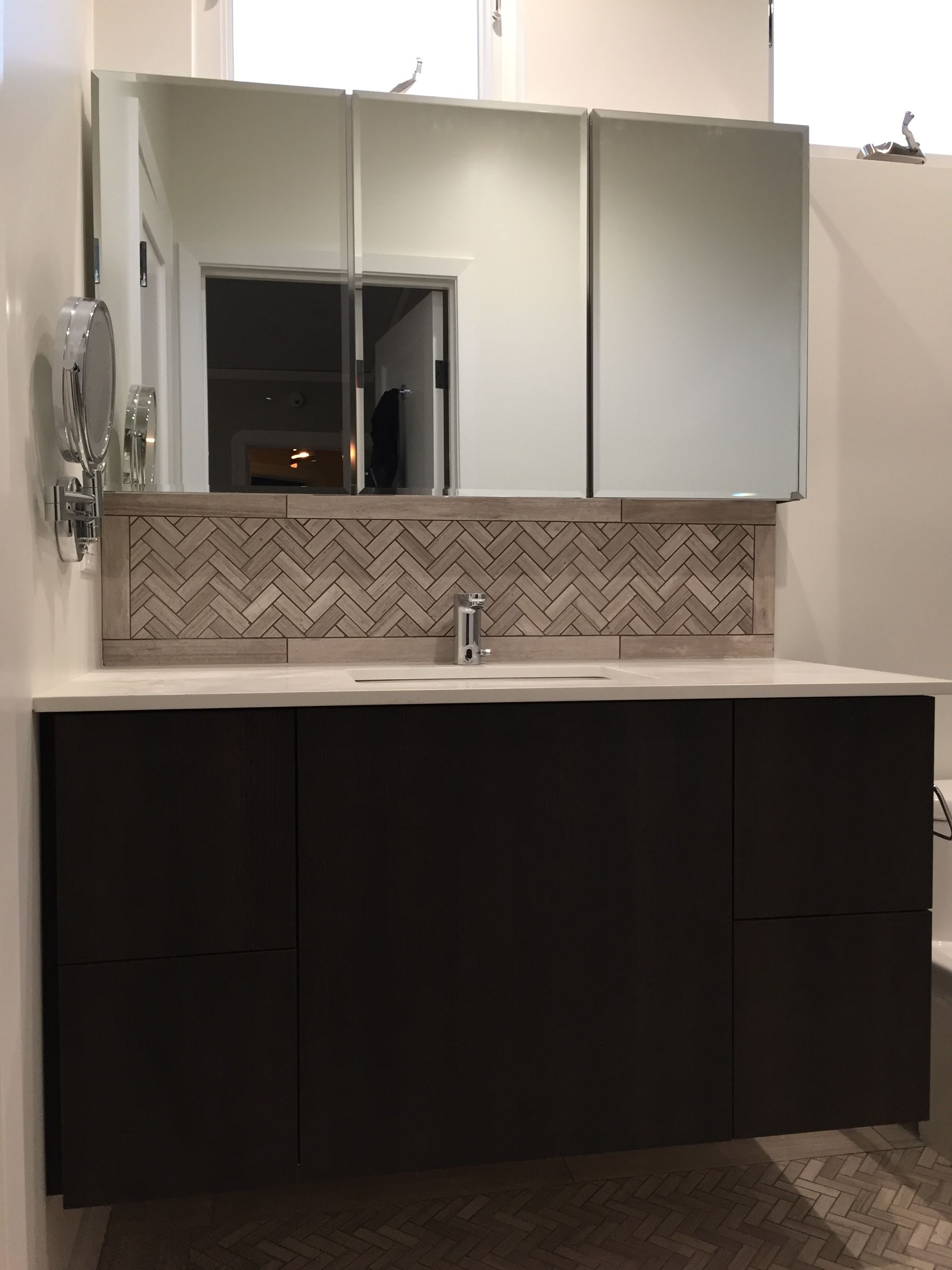 Modern bathroom vanity with dark cabinets, white countertop, mirrored cabinets, and herringbone tile backsplash