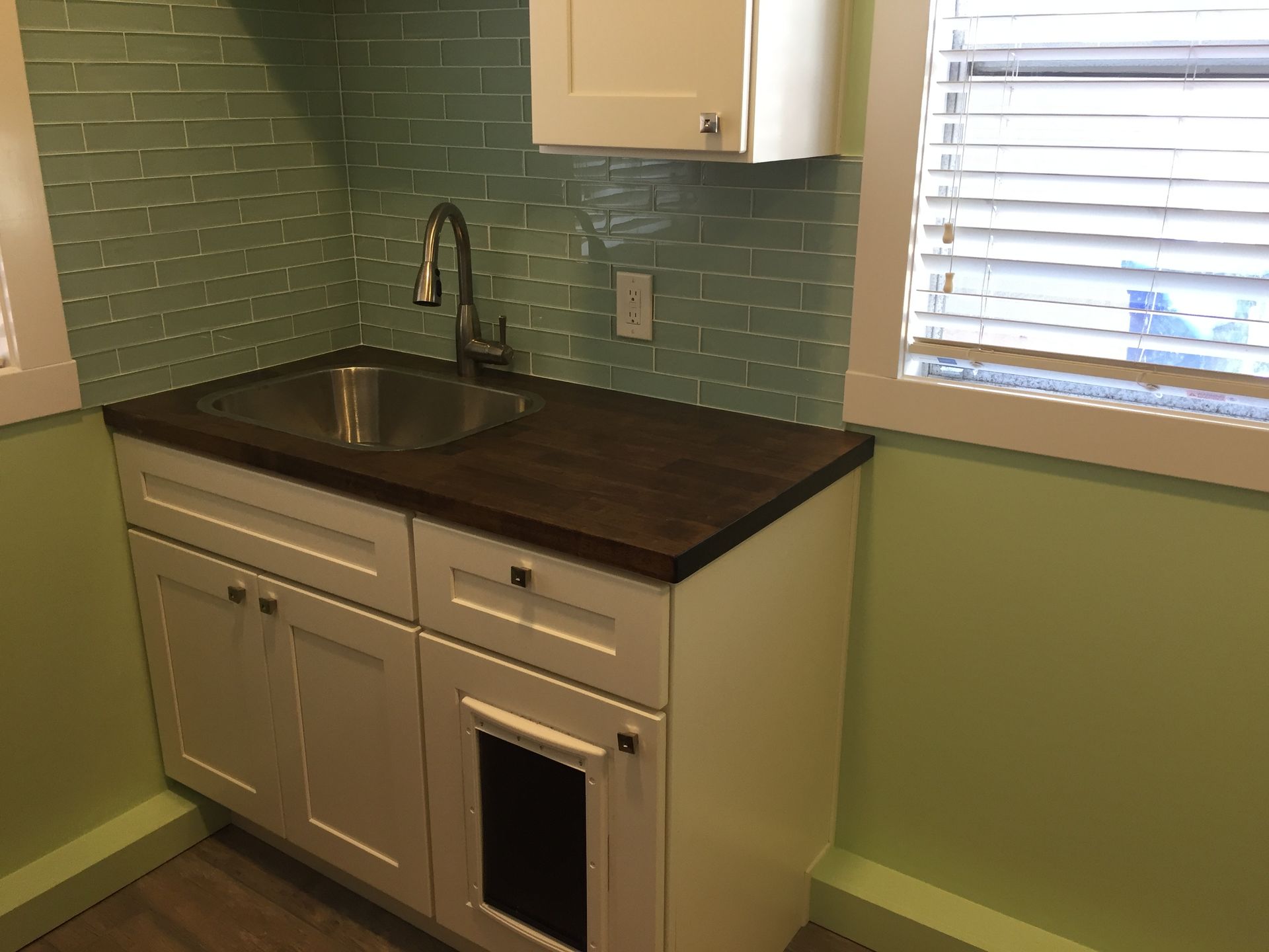Small kitchen corner with white cabinets, dark countertop, stainless sink, green tile backsplash, and window blinds.