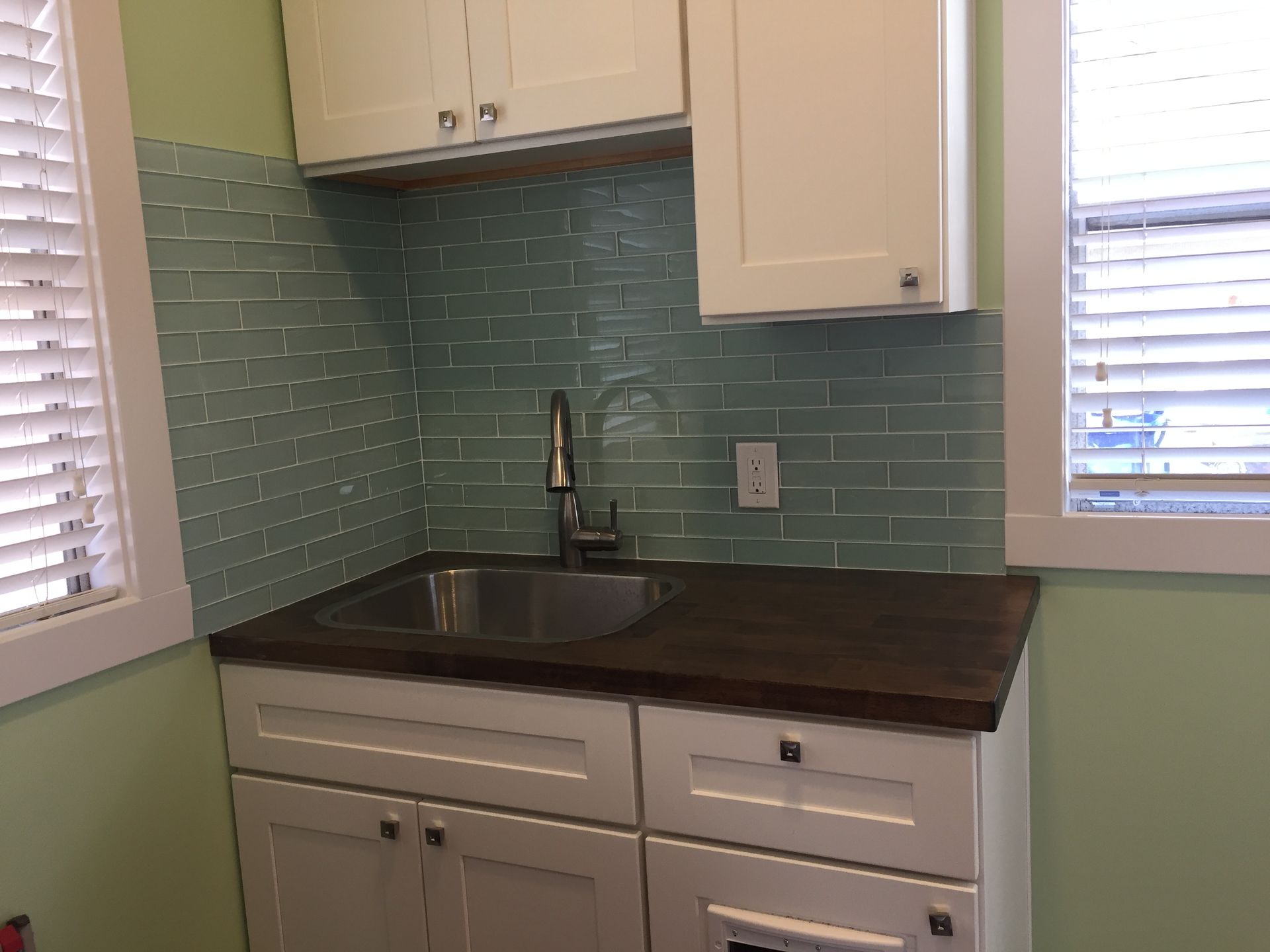 Small kitchen corner with mint tile backsplash, dark countertop, white cabinets, and a sink under cabinets and windows.
