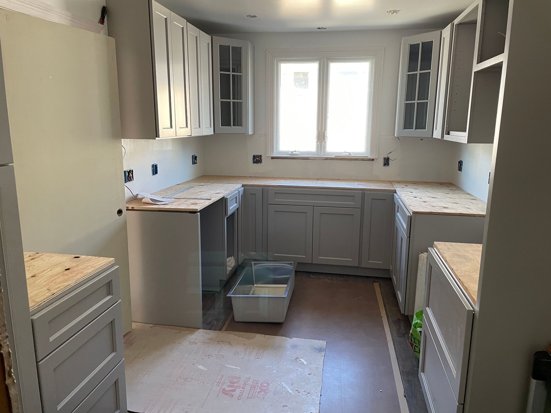 Empty kitchen with gray cabinets, white countertops, and a window over the sink under renovation.