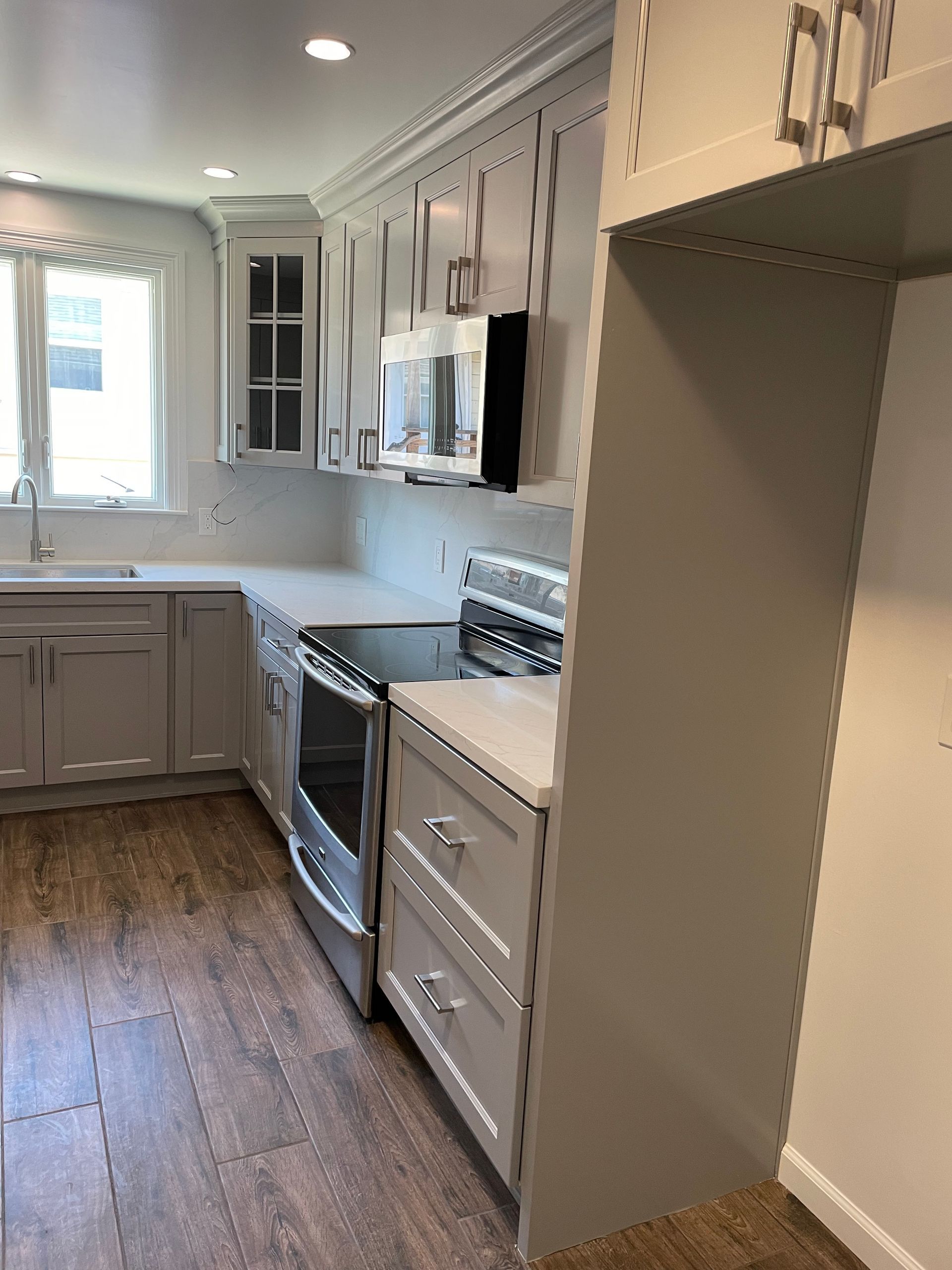 Bright white galley kitchen with gray cabinets, stainless stove, wood floors, and a window at the end.