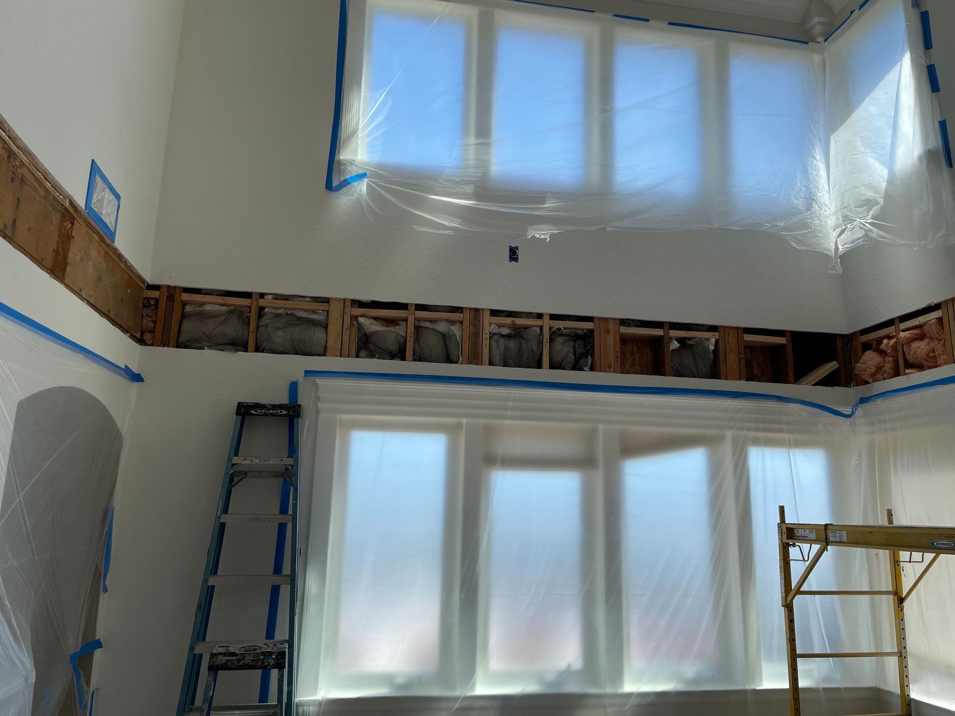 Interior room under renovation with covered windows, ladders, and exposed wall framing.