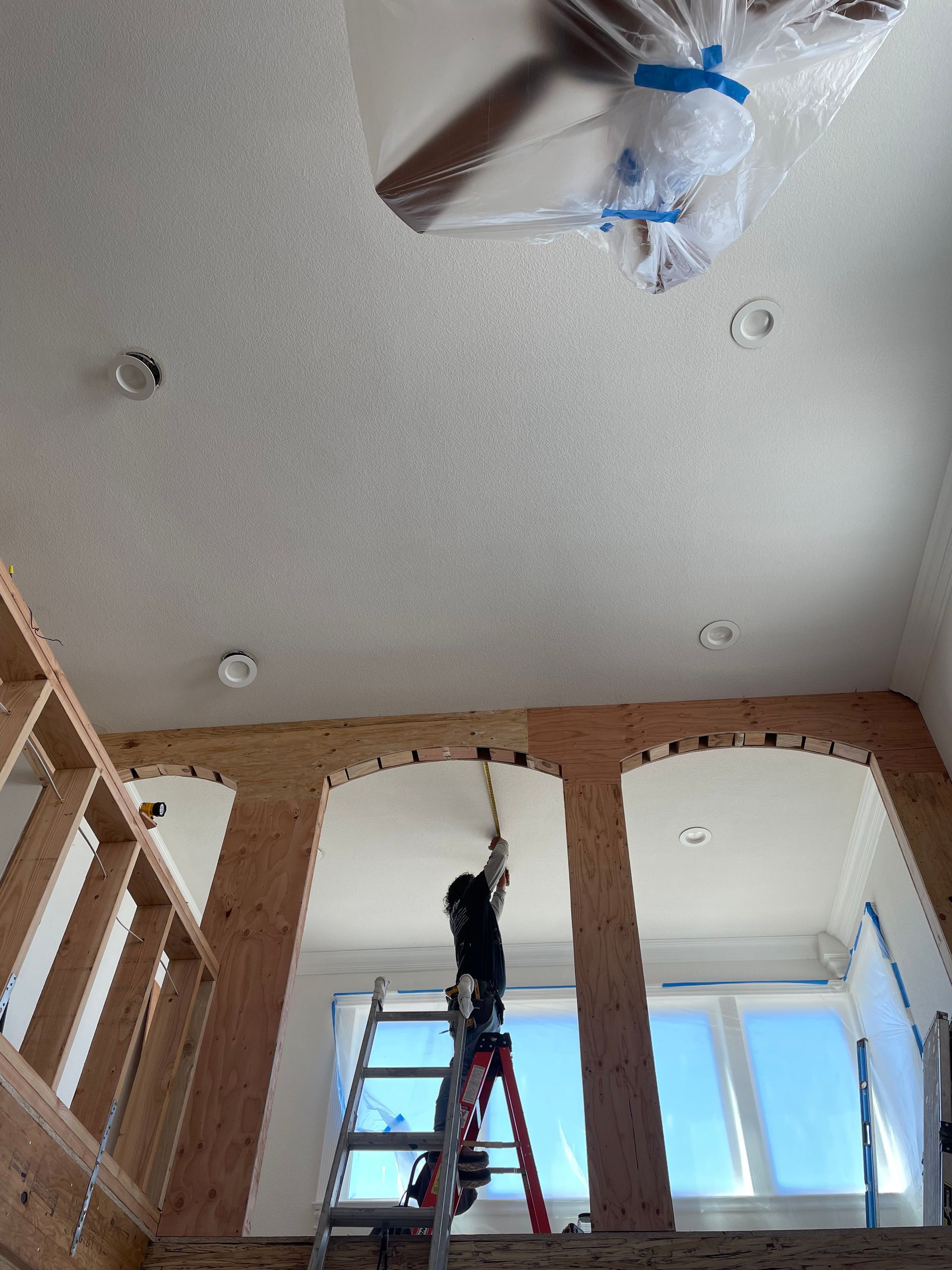Worker on a ladder installing a ceiling fixture in a room with unfinished walls and windows.
