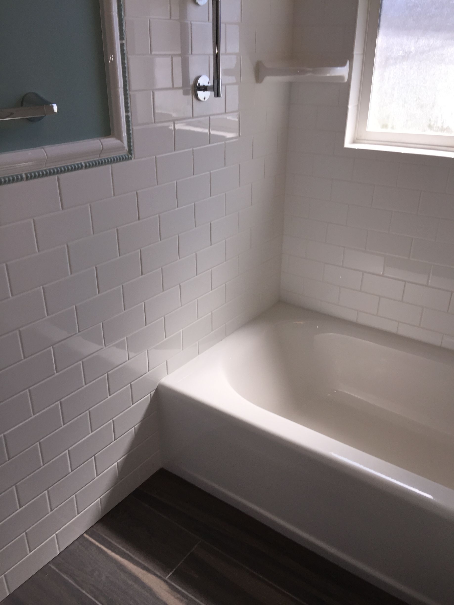 White-tiled bathroom corner with a bathtub, window, and dark wood-look floor.