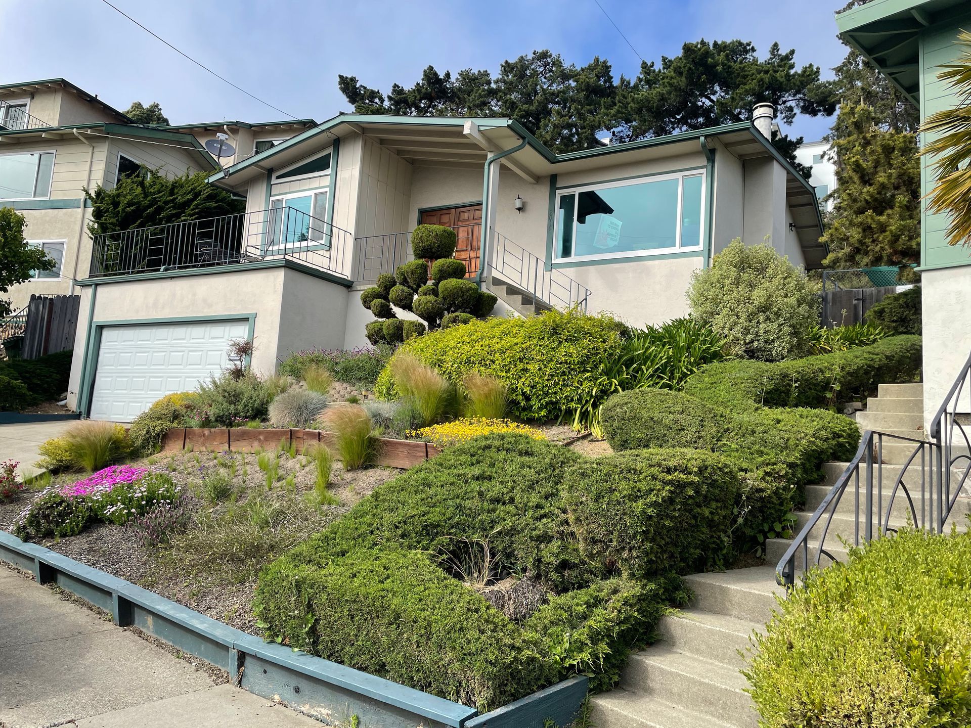 House on a steep hillside with terraced gardens, stairs, and a garage under a clear blue sky
