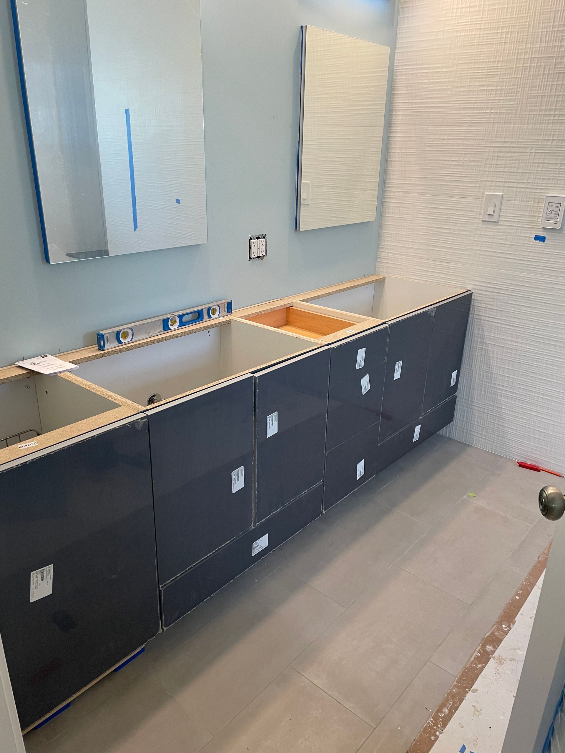 Bathroom under construction with dark cabinets, sink cutouts, and mirrors mounted on a white wall.