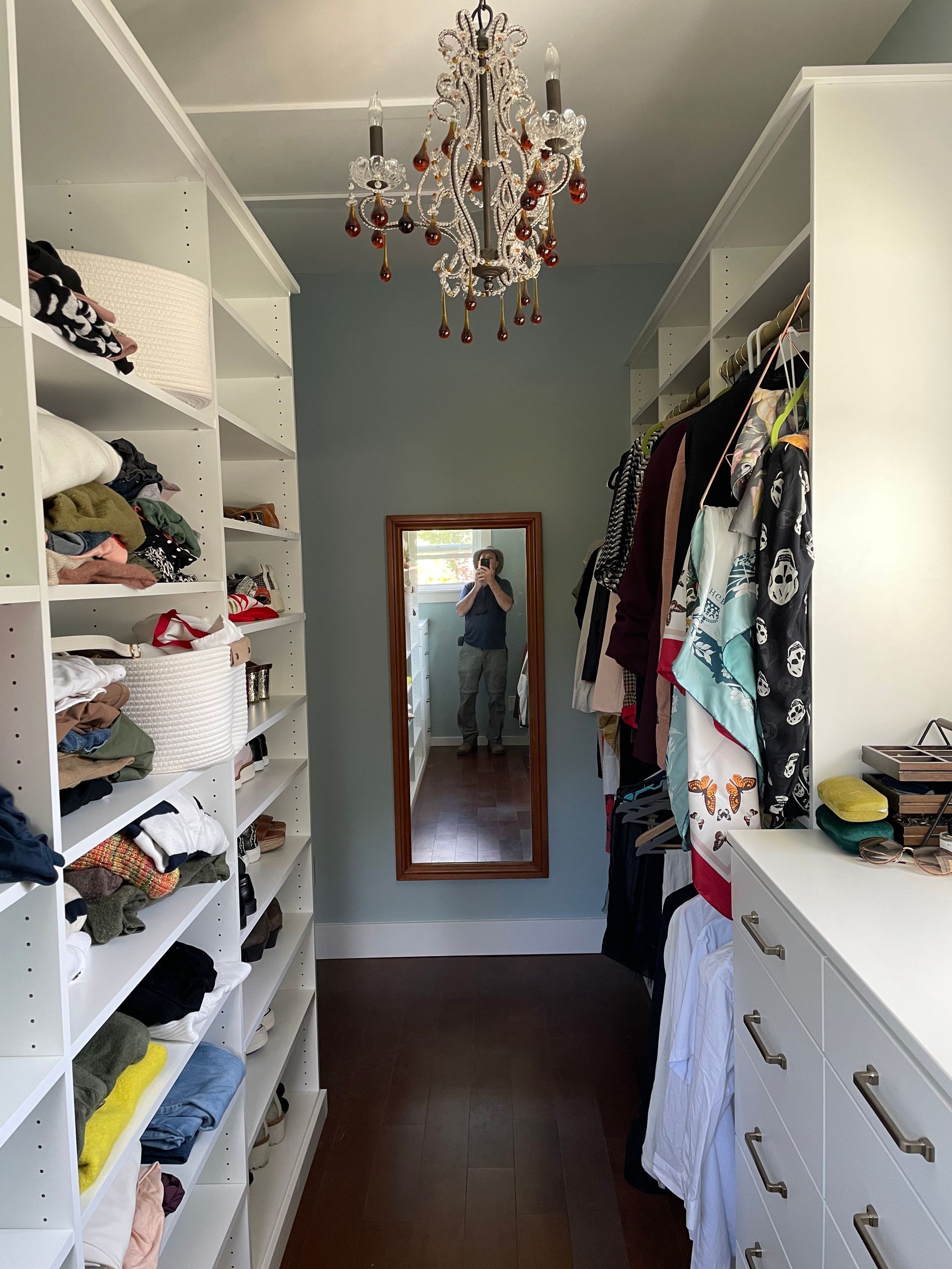 Walk-in closet with white shelves, hanging clothes, and a mirrored door reflecting the room.