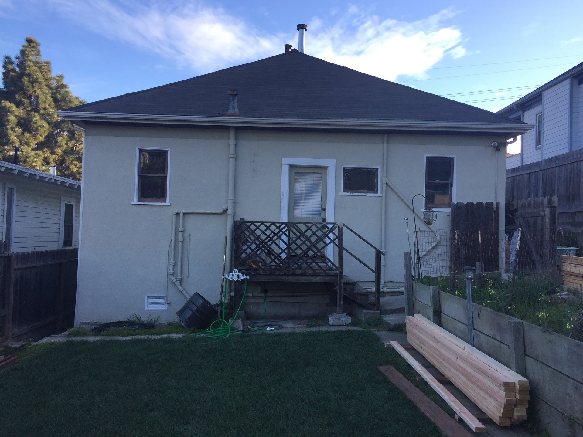 Backyard view of a small white house with a deck, lawn, and stacked lumber beside it.