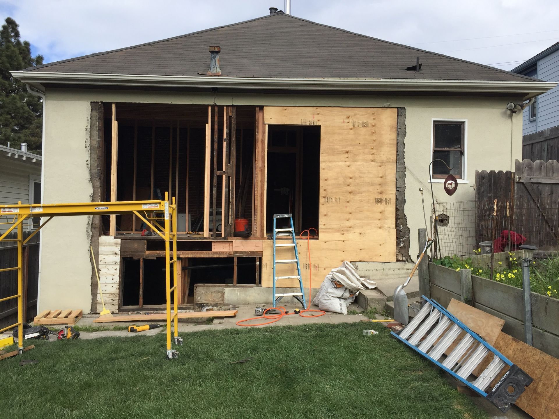 House exterior under renovation with open wall framing, scaffolding, ladder, and construction materials in a backyard.
