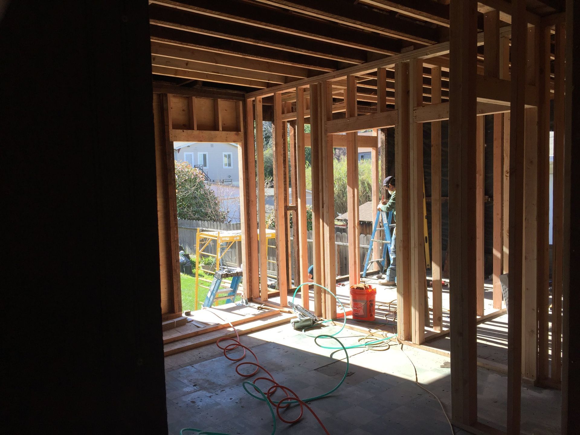 Unfinished house interior with exposed wooden framing, sunlight, tools, and a worker visible near the opening.