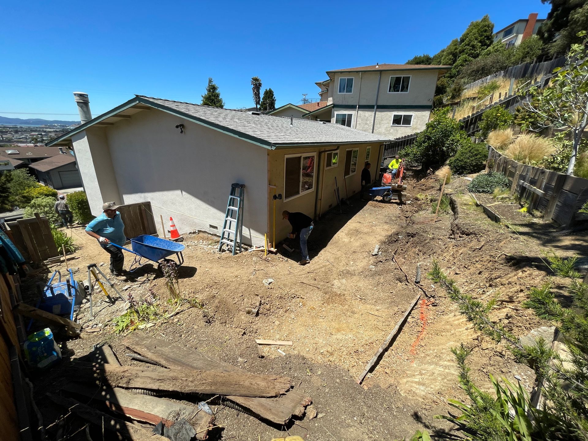 Backyard excavation beside a beige house, with dirt piles, tools, and workers near a hillside home