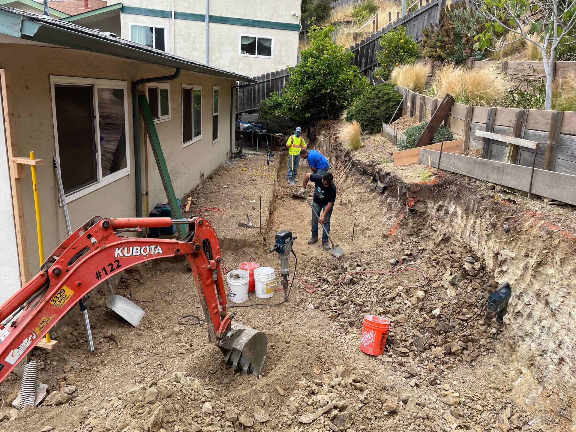 Backyard excavation with mini excavator and two workers beside a house and retaining wall