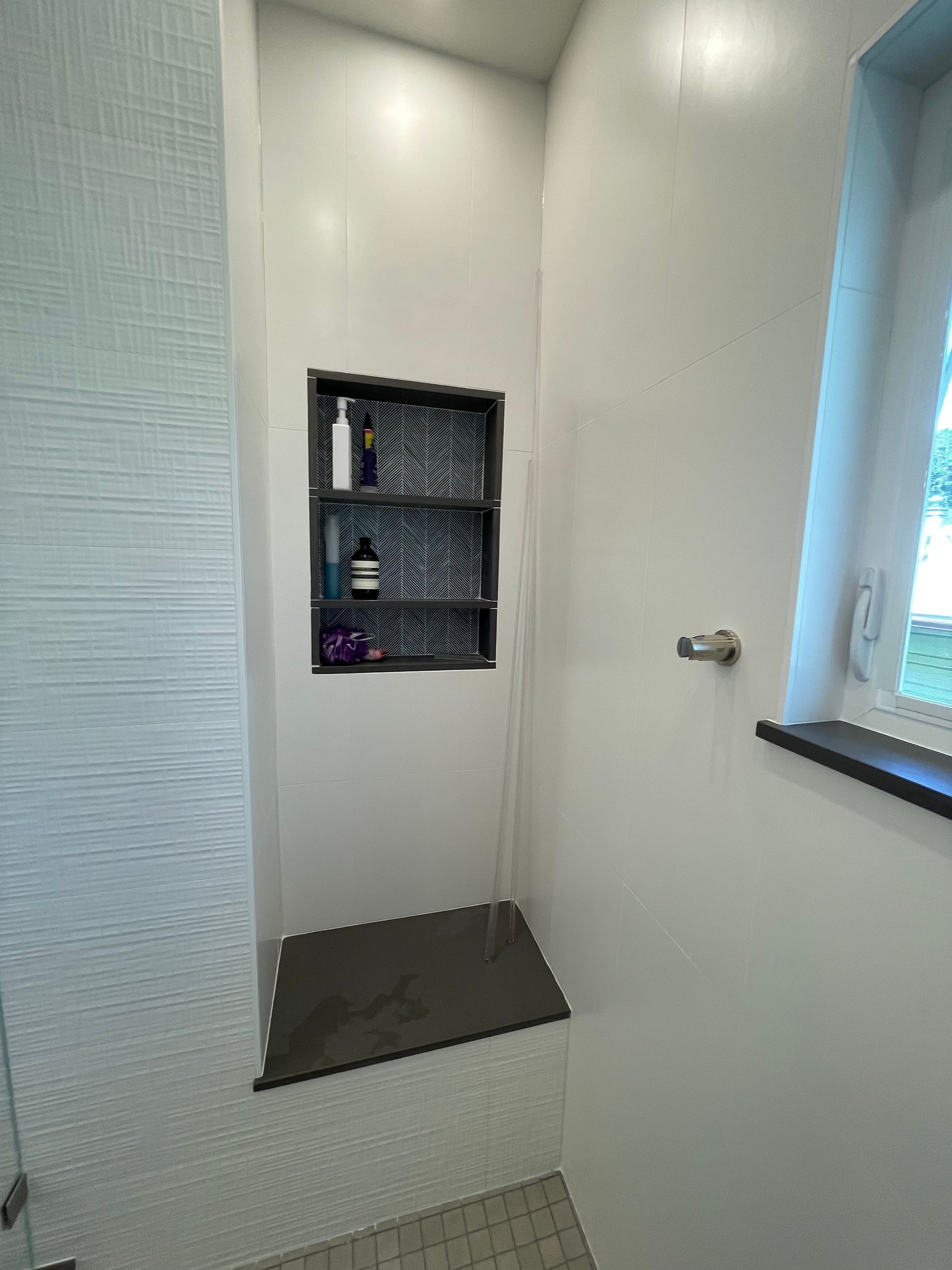 Built-in shower nook with gray tile shelf, dark floor ledge, and window light in a white bathroom