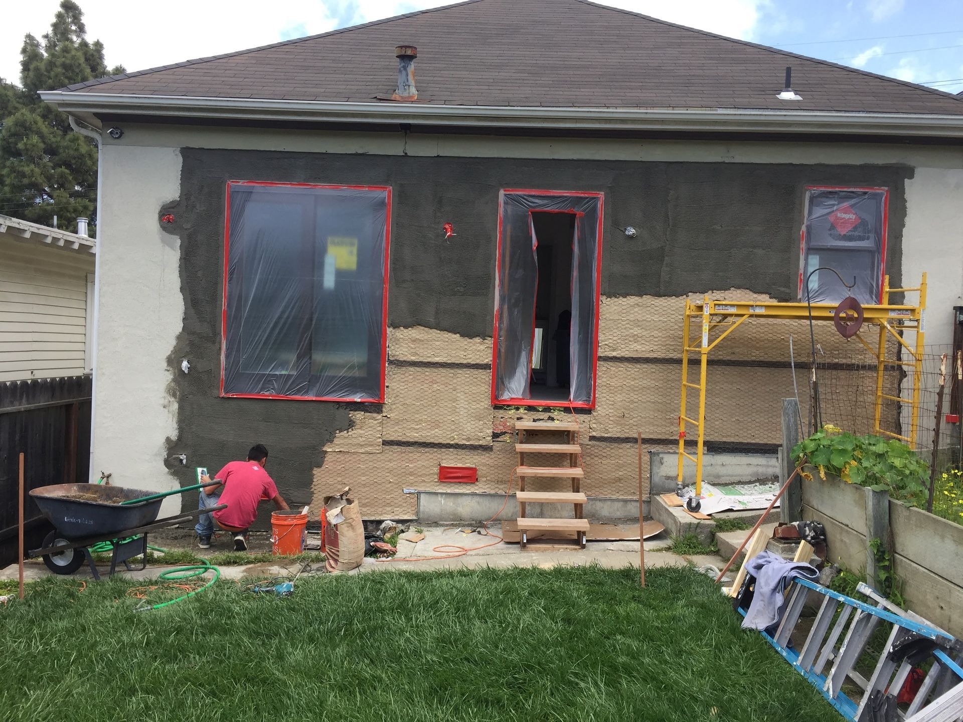 House exterior under renovation with two people working, covered windows, and ladders and tools in front
