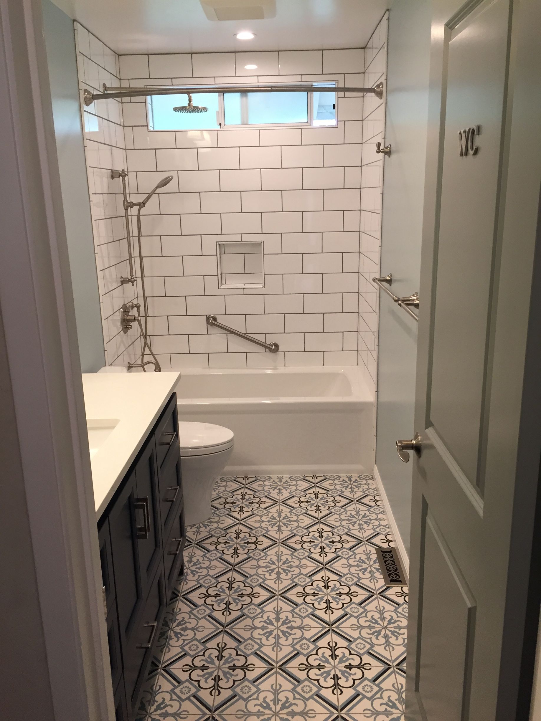 Narrow white bathroom with patterned tile floor, bathtub shower, toilet, vanity, and frosted glass door.