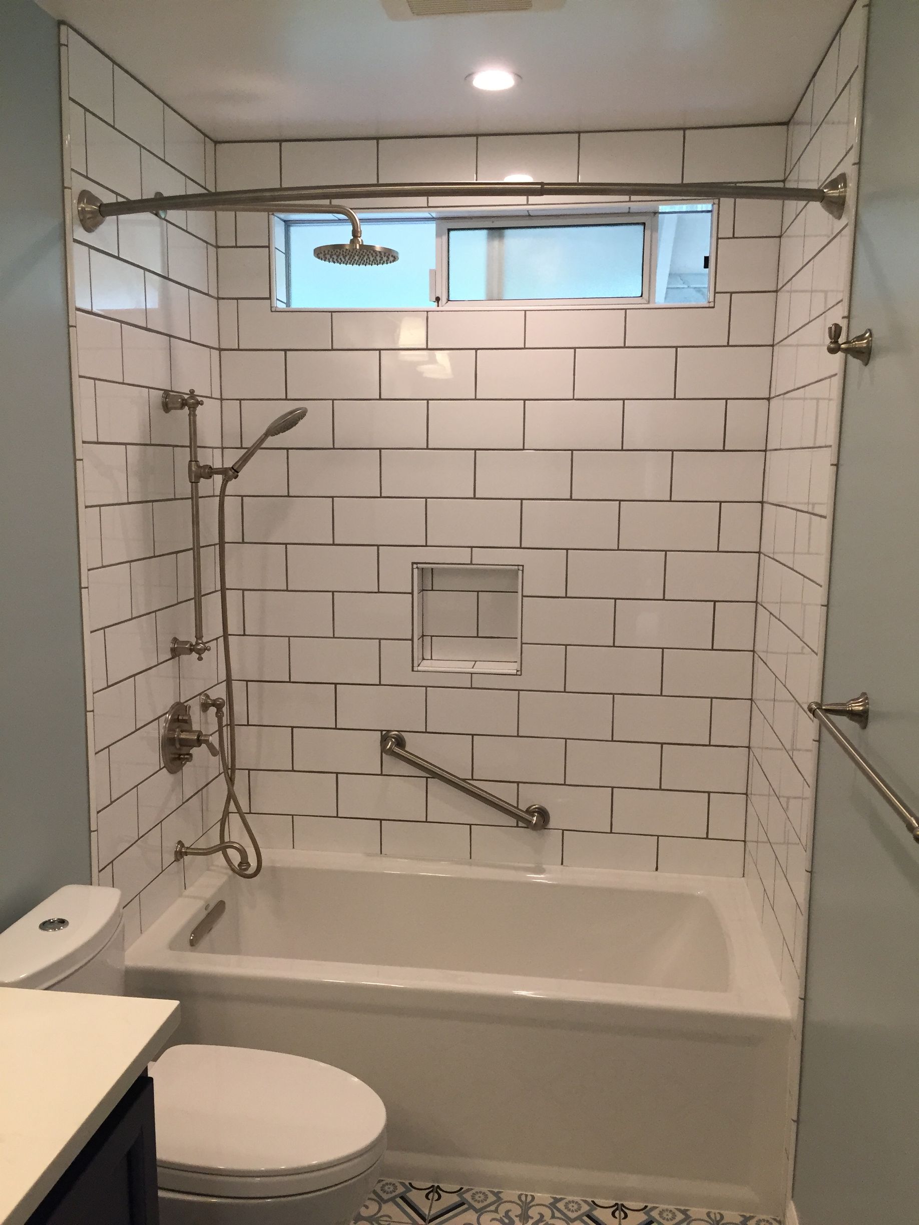 Accessible tiled bathroom with tub, grab bars, handheld shower, and toilet under a small window