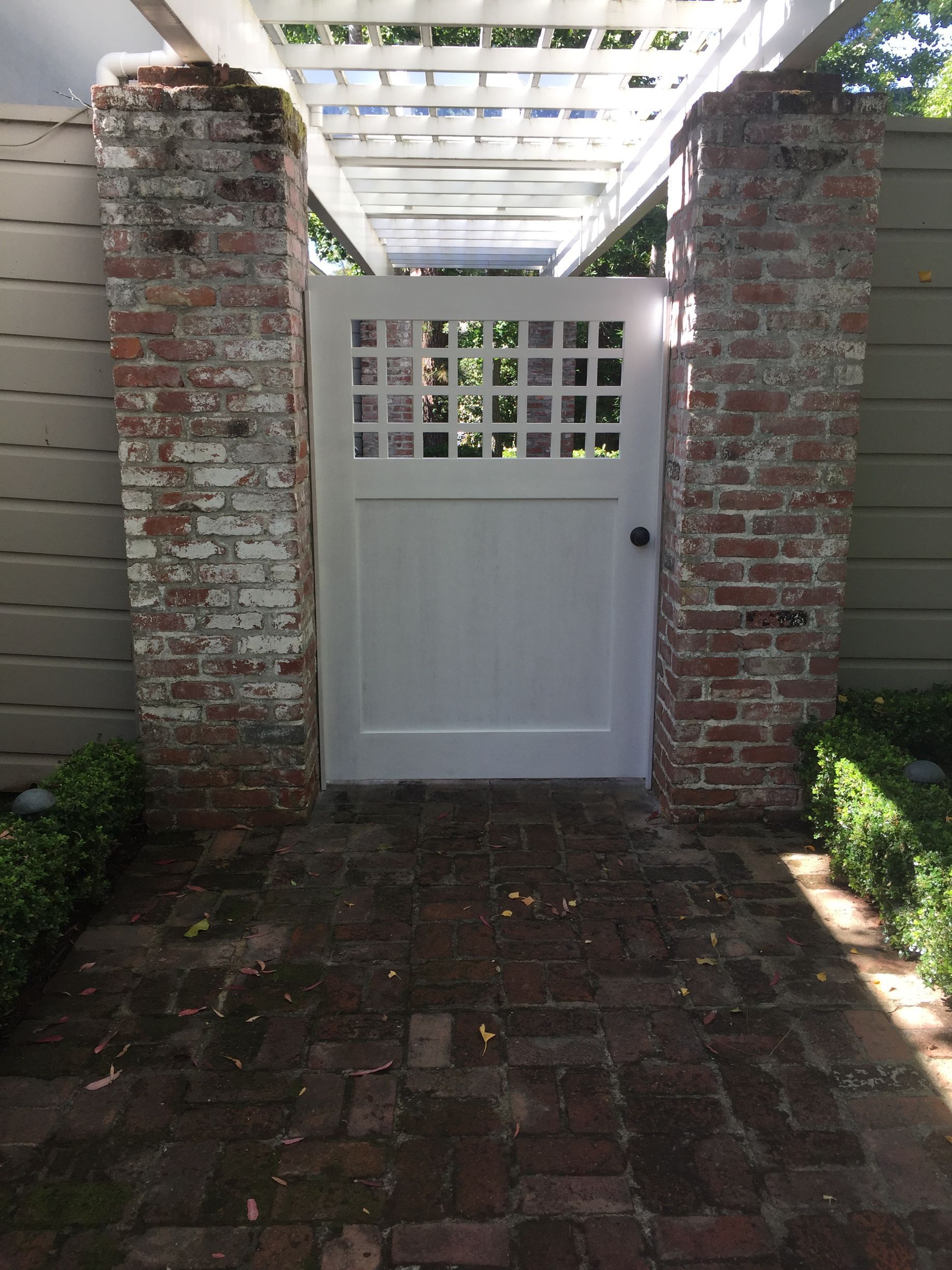 White gate set between brick pillars in a brick walkway under a pergola