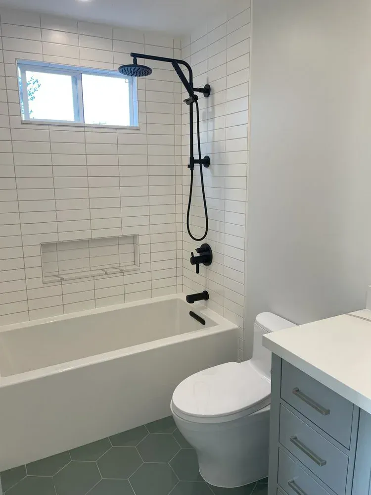 Modern bathroom with white tile, bathtub, shower, toilet, and vanity near a small window.
