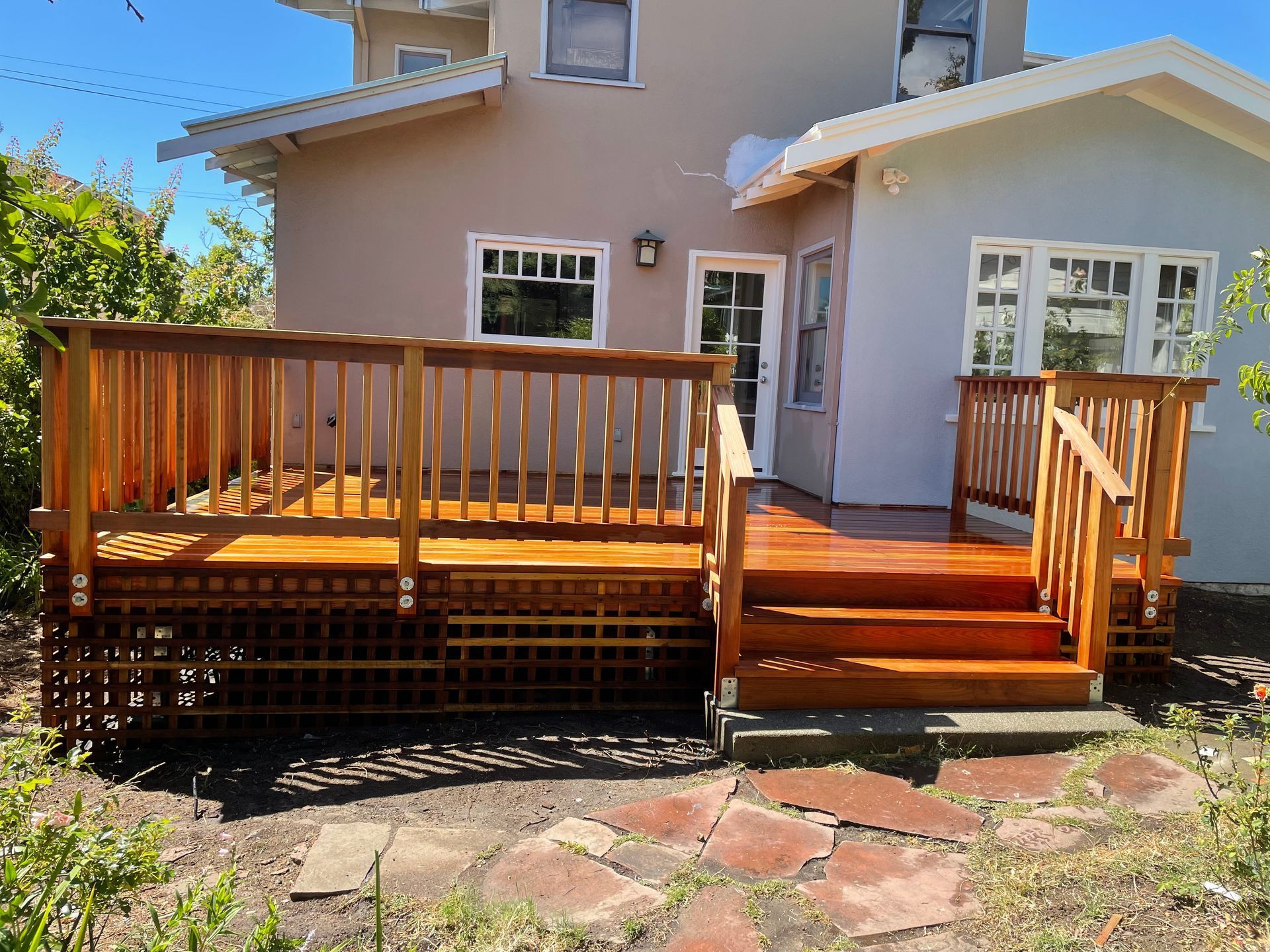 A newly stained wooden deck with railings and steps attached to the rear of a multi-story, light-colored house.