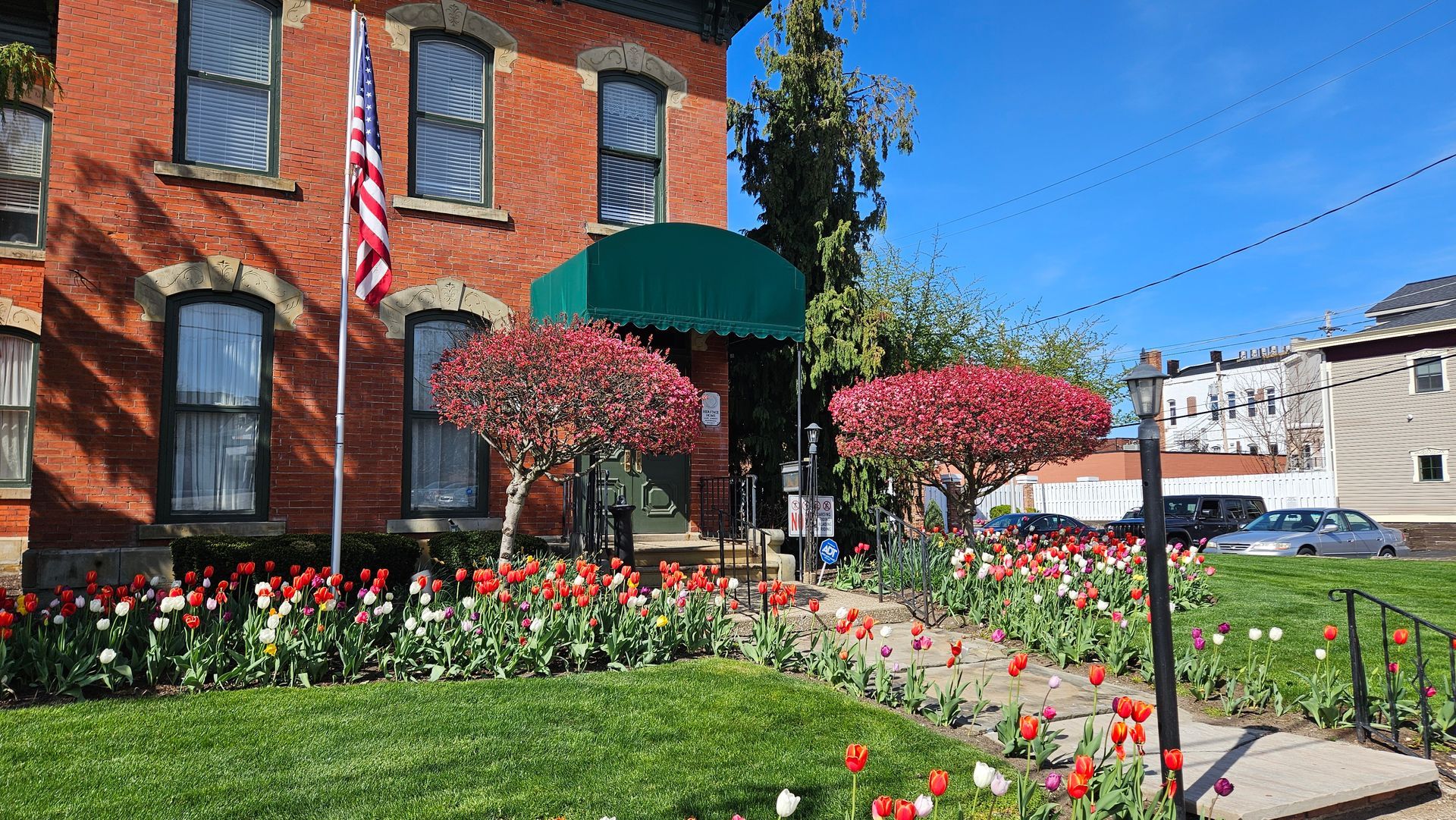 Brick building with green awning, American flag, tulip-lined walkway, flowering trees, and a clear blue sky.