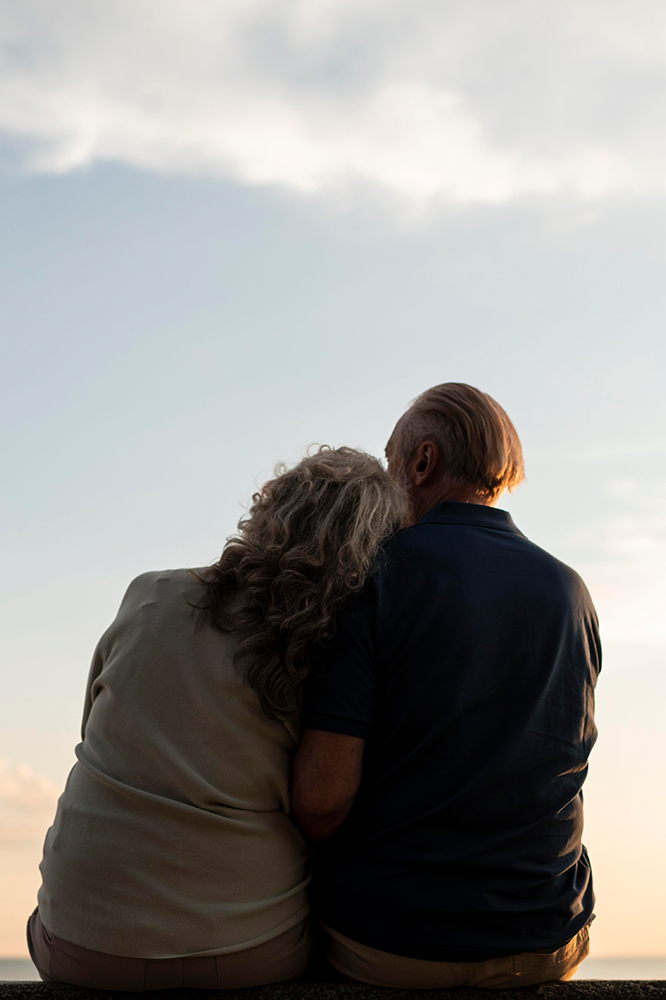 An older couple sits close, looking at a bright sky. Woman rests head on man's shoulder.