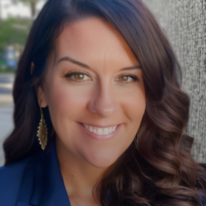 Woman with brown hair smiles, wearing a navy blazer and gold earrings.