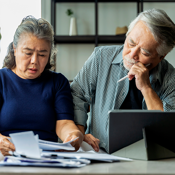 Older couple reviewing documents at a table; concerned expressions, tablet present.