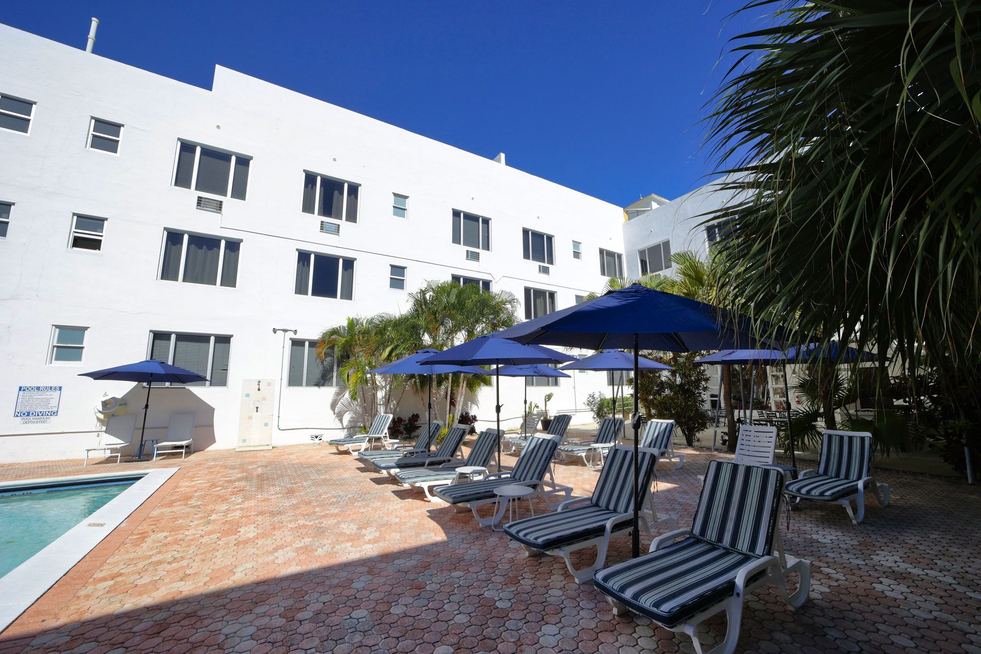 Poolside patio with lounge chairs and blue umbrellas beside a white hotel building
