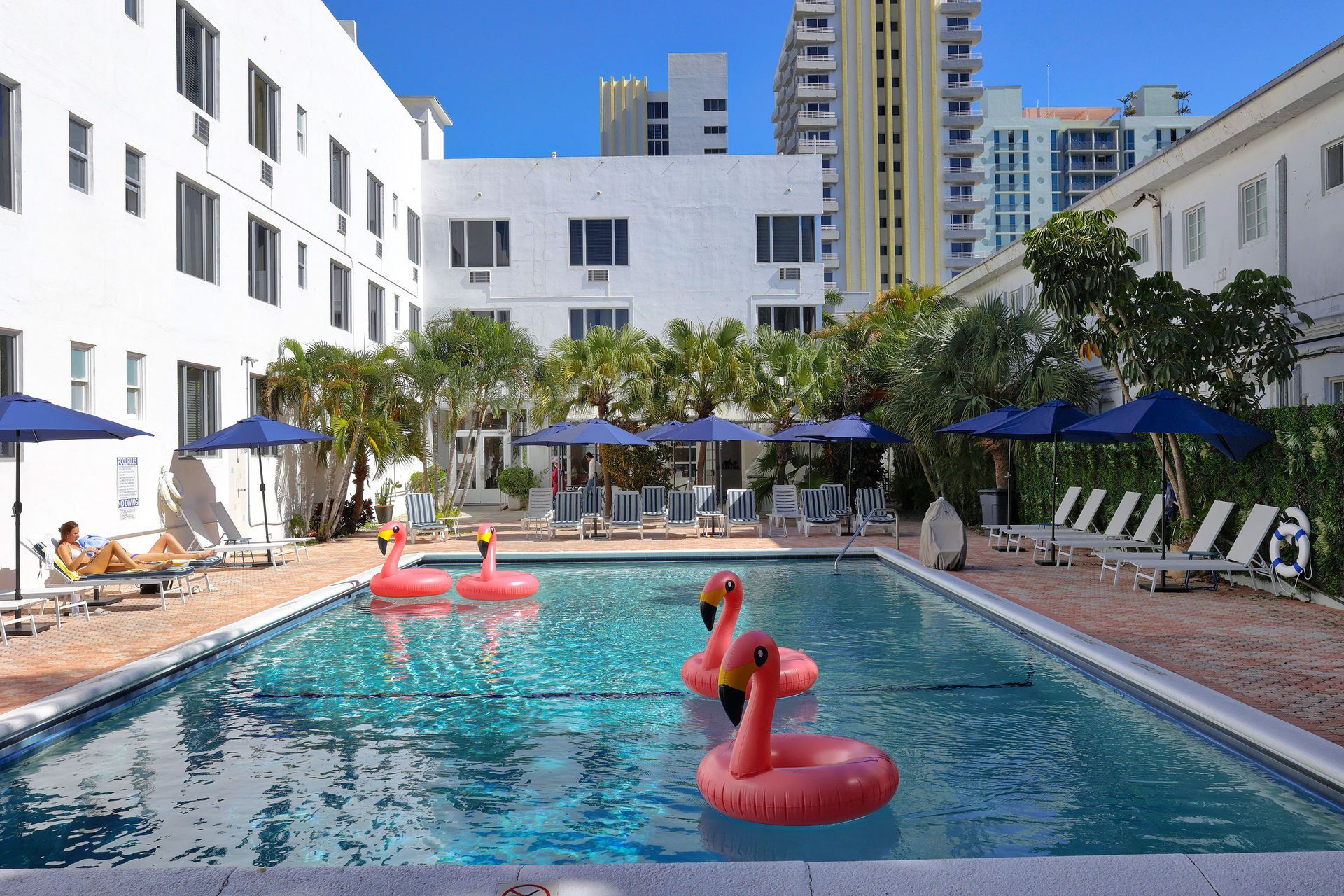 Outdoor pool with pink flamingo floats, blue umbrellas, and white apartment buildings in the background