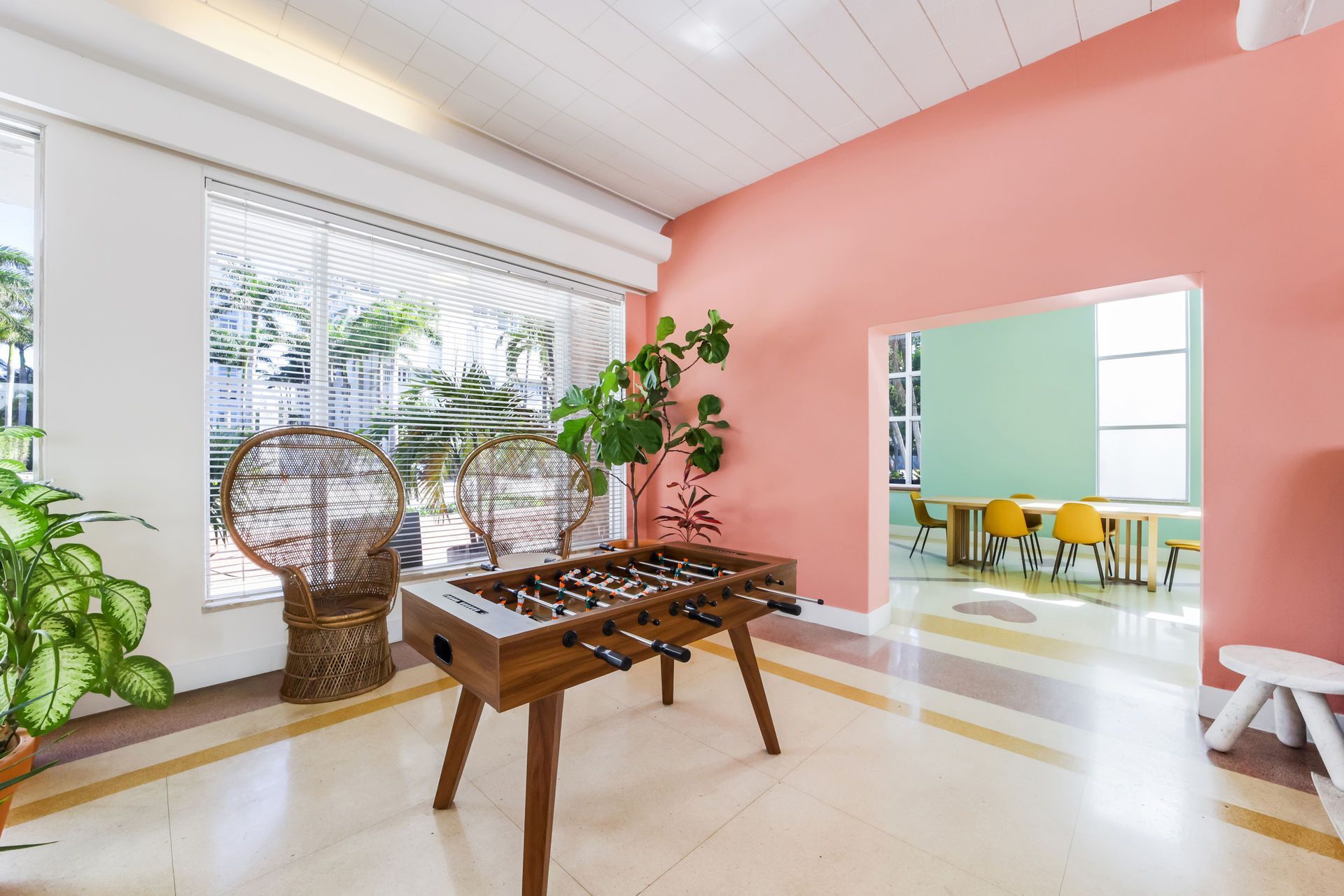 Bright sunroom with a foosball table, wicker chair, pink wall, and open view into a dining area