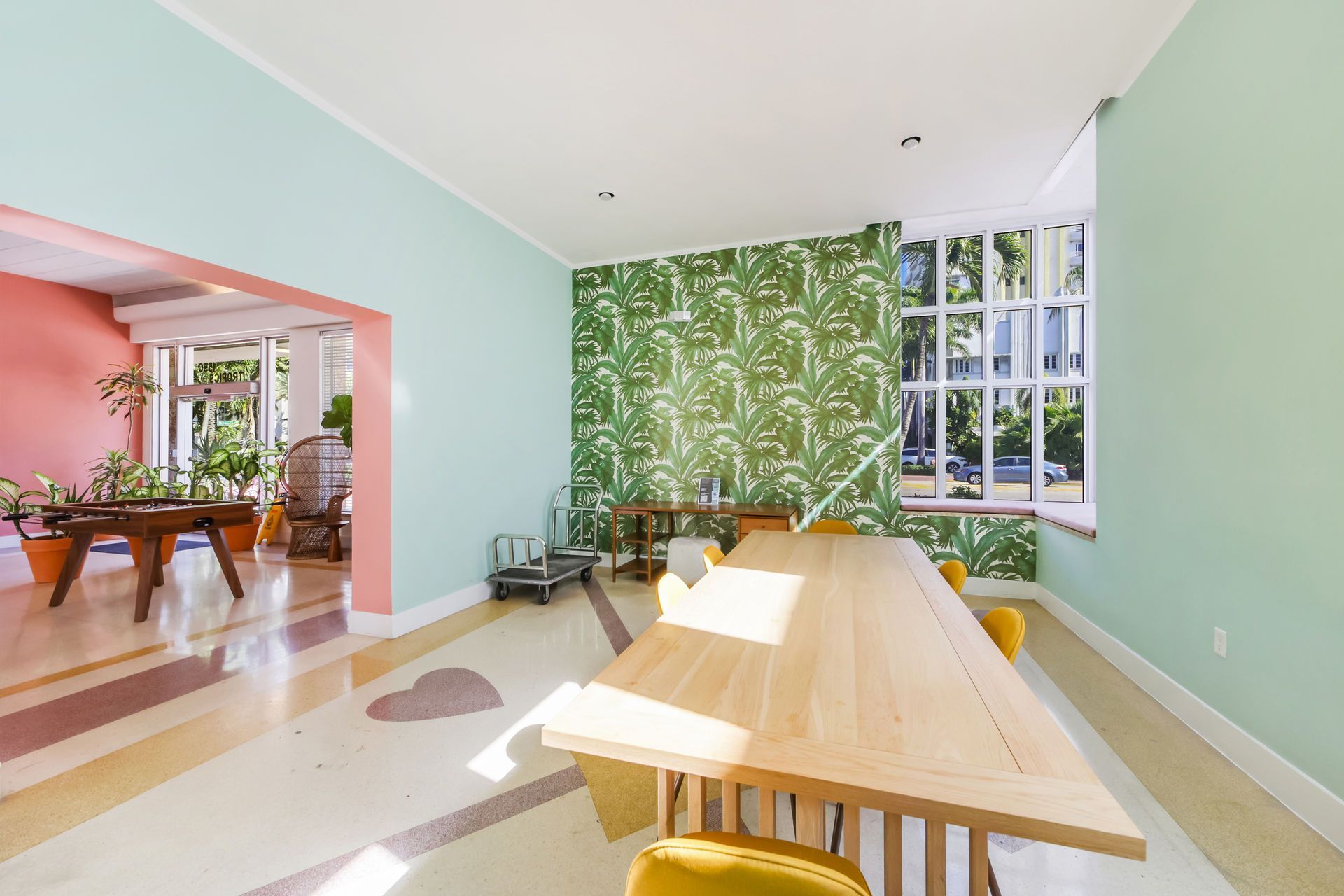 Bright dining room with a wooden table, mint walls, pink adjoining room, and leafy wallpaper accent wall