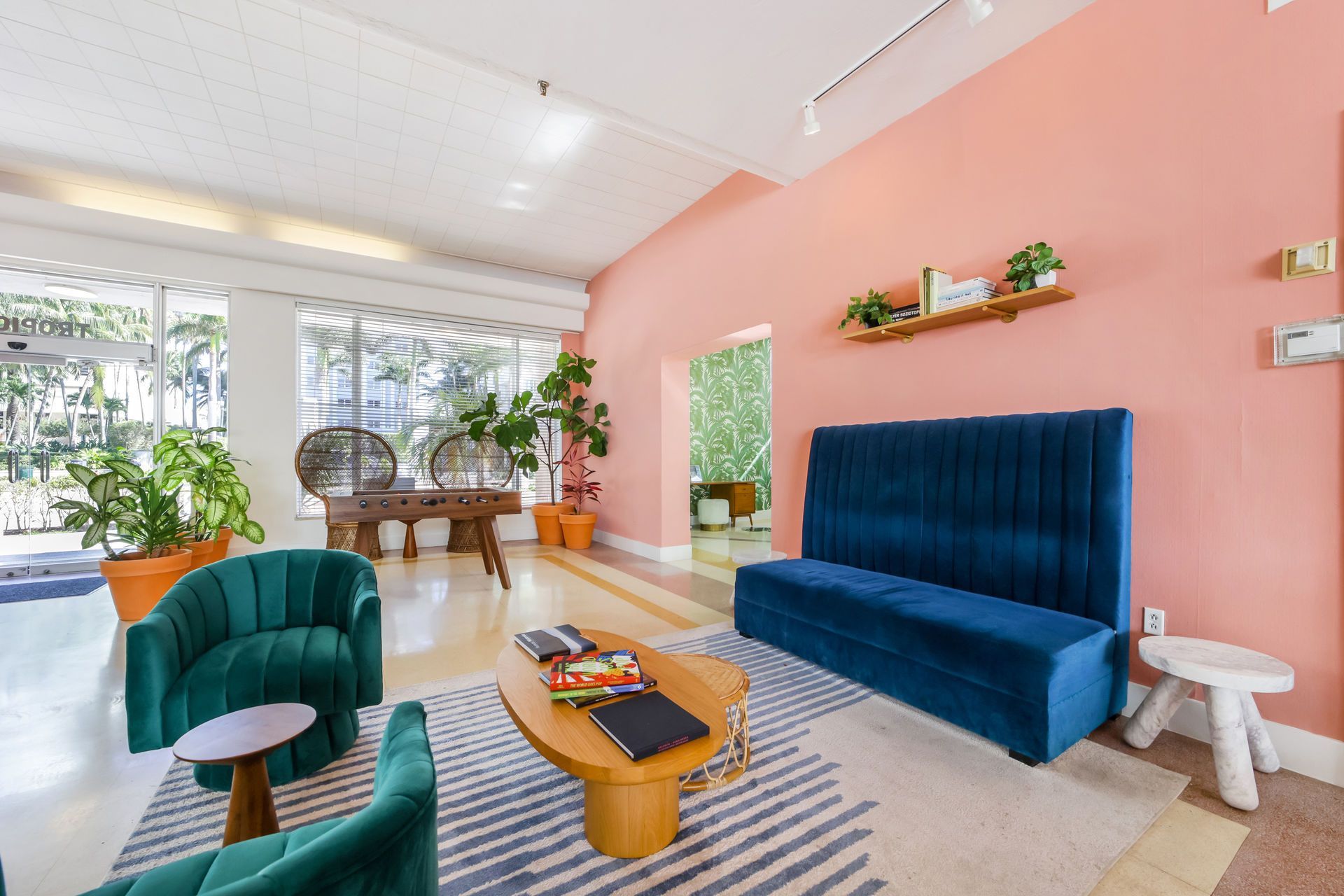 Bright living room with teal chair, blue sofa, pink walls, plants, and sunlight from large windows
