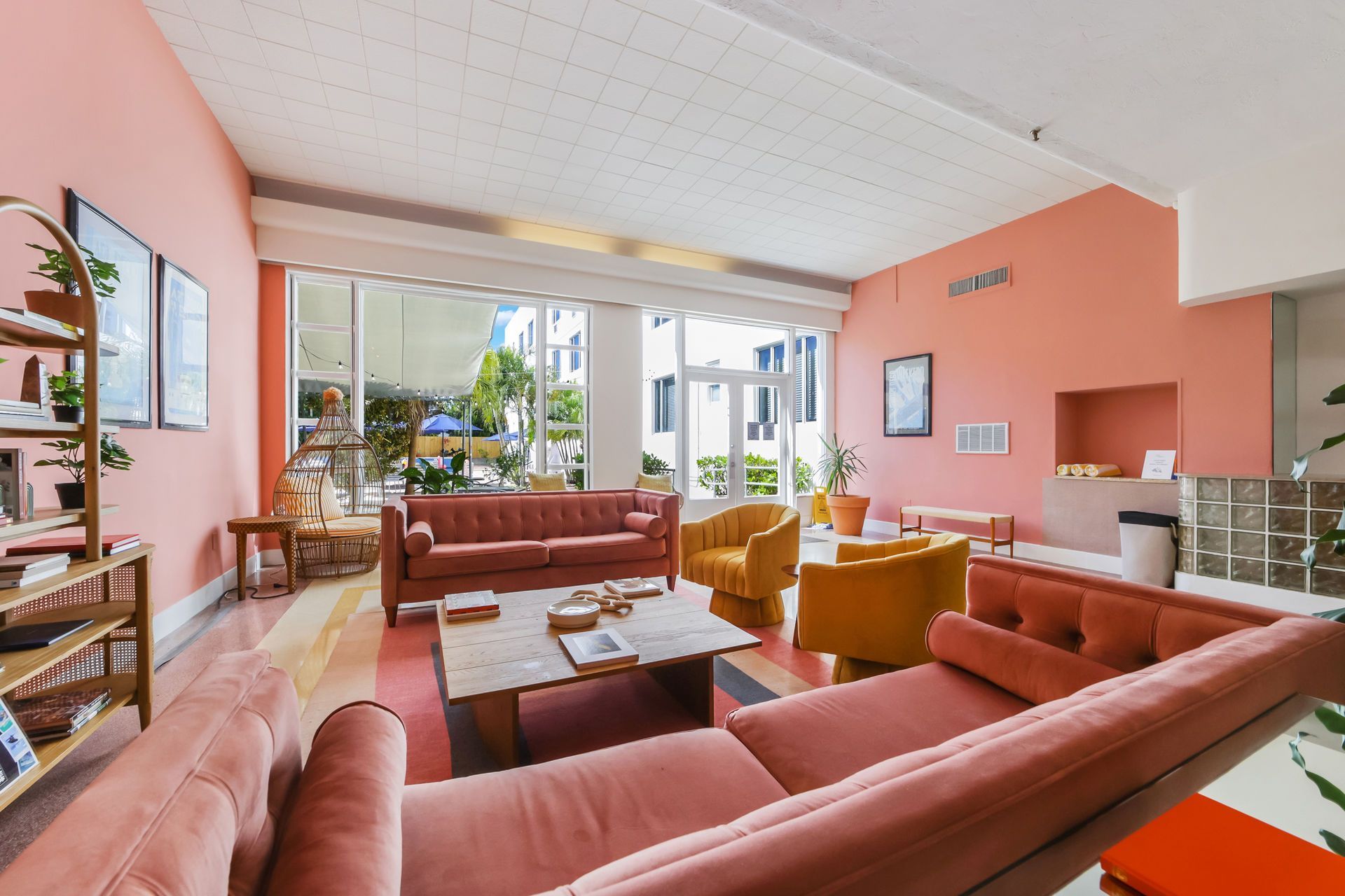 Bright living room with pink walls, coral sofas, mustard chairs, and large windows overlooking a patio.