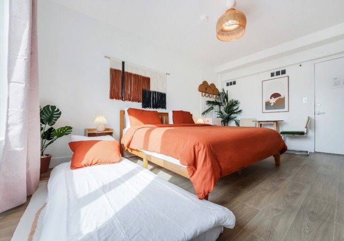 Bright minimalist bedroom with orange bedding, white walls, and a cushioned chair by the window