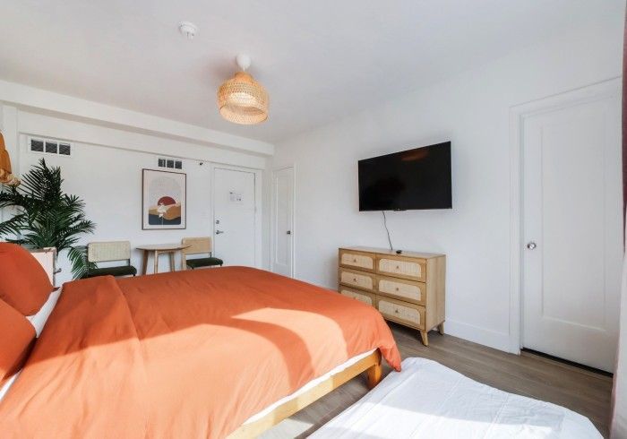 Bright bedroom with orange bedspread, wall-mounted TV, dresser, and white walls.