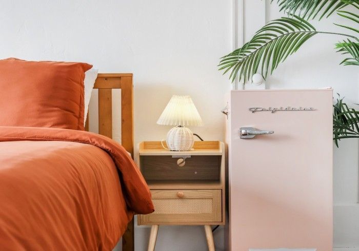 Cozy bedroom corner with orange bed, wooden nightstand, lamp, and pale pink mini fridge