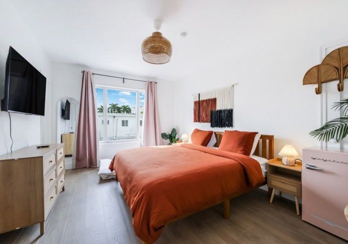 Bright bedroom with orange bedspread, white walls, TV, dresser, and balcony doors with pink curtains
