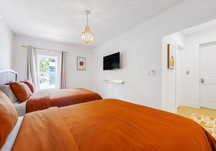 Bright white bedroom with orange bedding, wall-mounted TV, window, and patterned chair
