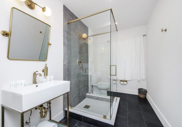Modern white bathroom with black tile floor, glass shower, wall-mounted sink, and gold fixtures