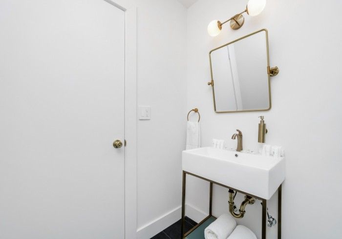 Minimal white bathroom vanity with gold mirror and wall light, black-framed sink stand, and closed door.