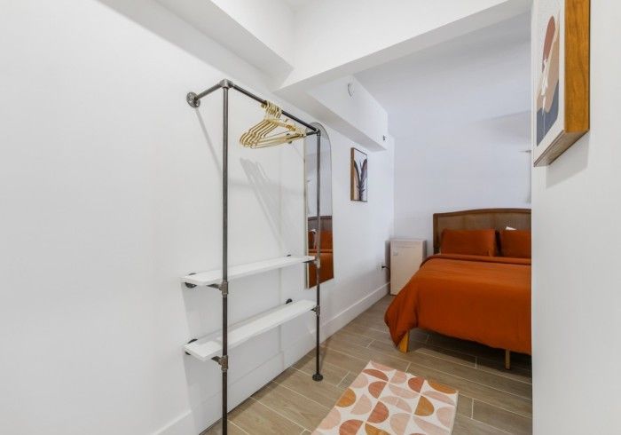 Small bedroom with metal clothing rack, white shelves, orange bed, and patterned rug.