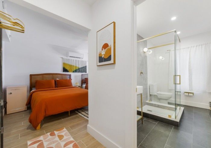 Bright bedroom with orange bedspread, white walls, and a glass shower room beside it.