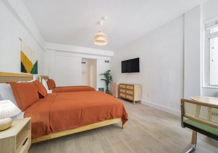 Bright bedroom with orange bed, wall-mounted TV, wicker chair, and large window